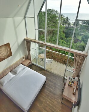 A bedroom with a large bed, a wooden desk, and a balcony with a view of the ocean and greenery. - Negla Beach Villa (Purbayani)