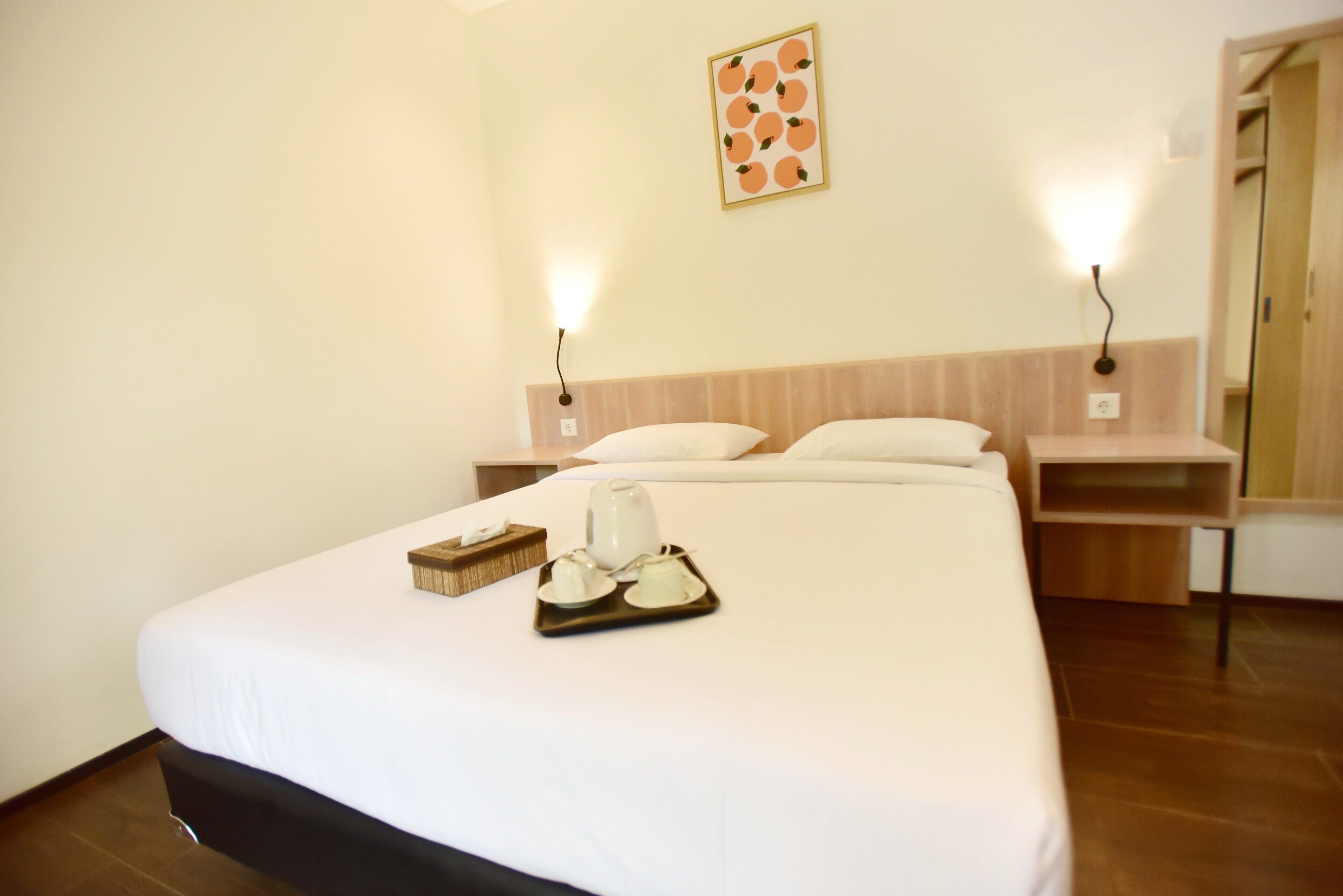 A hotel room with a large bed, a tray with tea and a teapot, two bedside lamps, and a fruit-themed wall decoration.