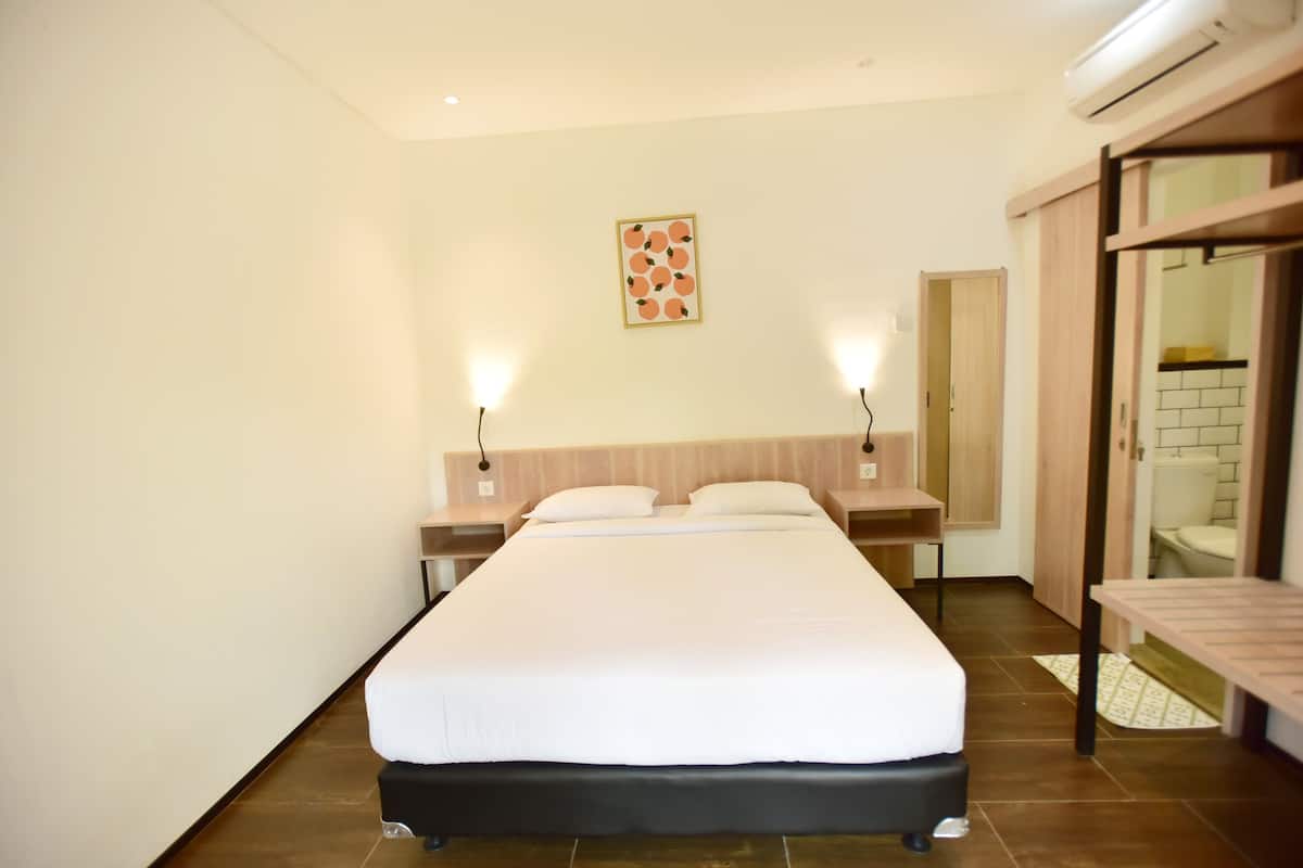 Habitación de hotel ordenada con una cama grande, dos mesitas de noche, una lámpara de pared y una puerta que da al baño.