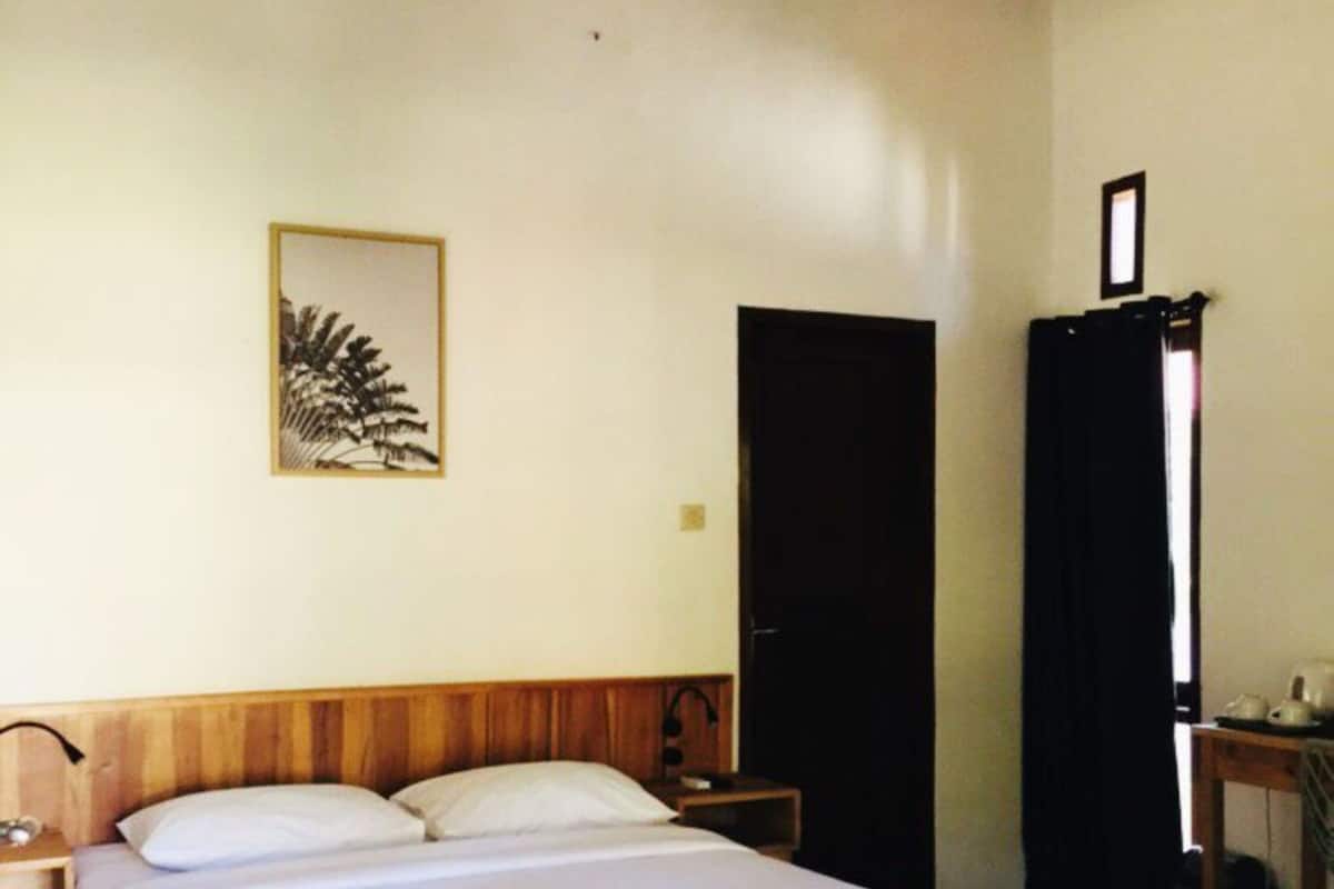 Una habitación con una cama grande, un cabecero de madera, una mesita de noche, un cuadro enmarcado en la pared, una puerta y una pequeña mesa con una lámpara.