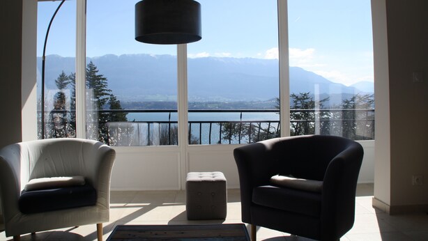 A room with two chairs, a small ottoman, and a hanging lamp. A large window offers a view of mountains and a lake.