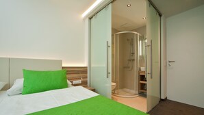 A modern hotel room with a bed, a green accent pillow, a glass-enclosed shower, and a wooden headboard. - Happy House das fröhliche Urlaubszuhause (Krumpendorf am Woerther See)