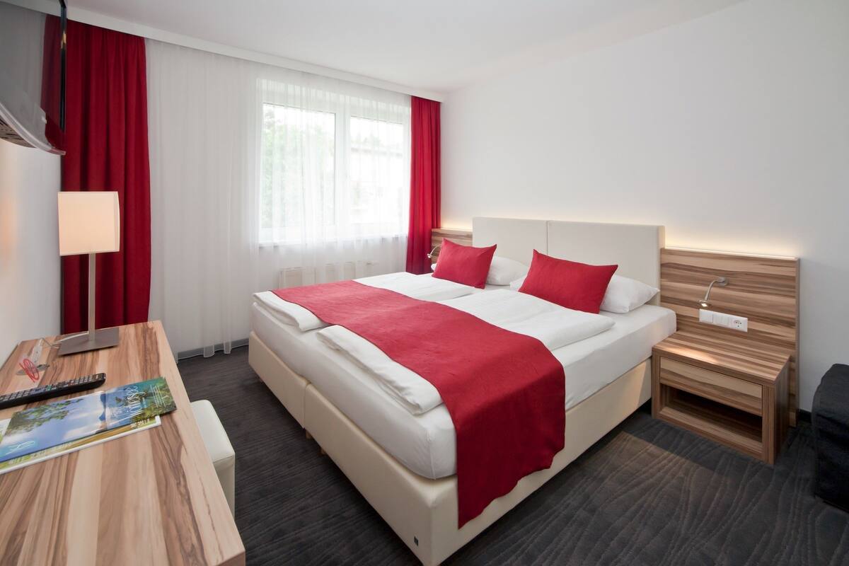 A hotel room with a large bed, red curtains, a desk with a lamp, and a view of the outdoors through the window.