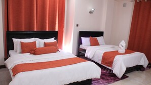 Habitación de hotel con dos camas, cada una con ropa blanca y detalles naranja, una alfombra púrpura y cortinas rojas.