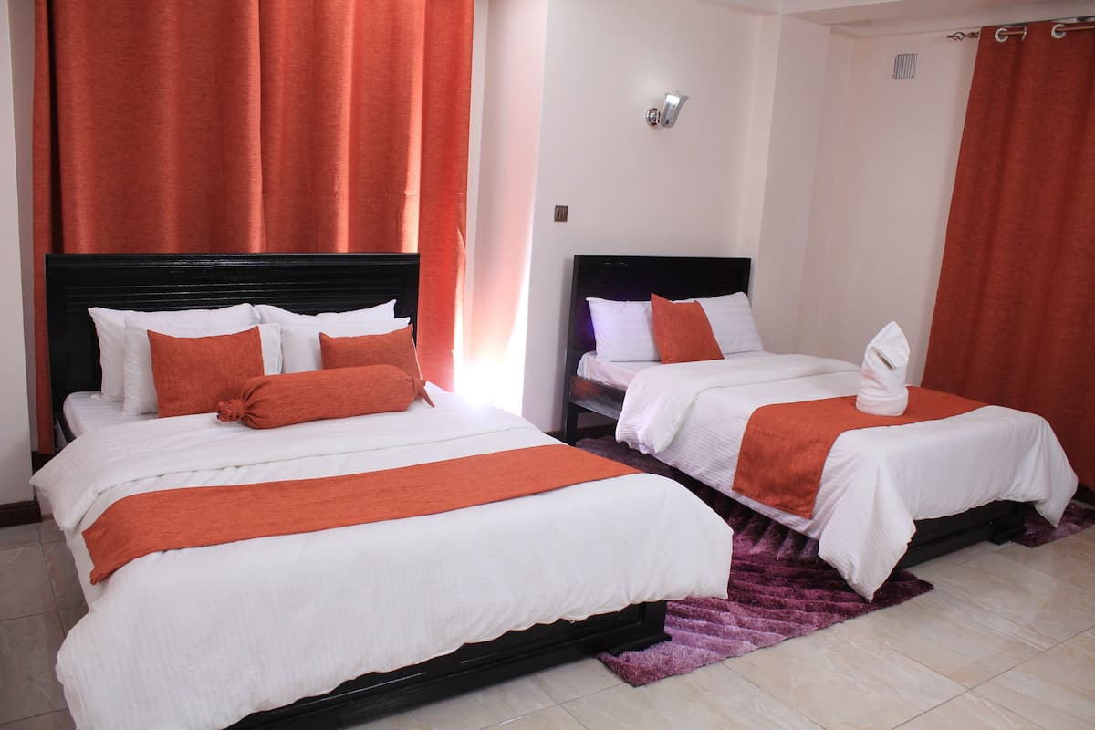 Habitación de hotel con dos camas, cada una con ropa blanca y detalles naranja, una alfombra púrpura y cortinas rojas.