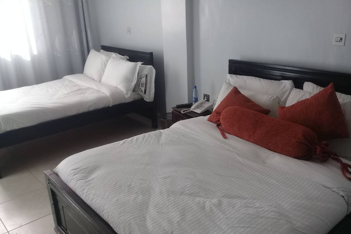 Dos camas individuales con ropa de cama blanca, una mesita de noche con un teléfono y una botella, y un enchufe de pared.