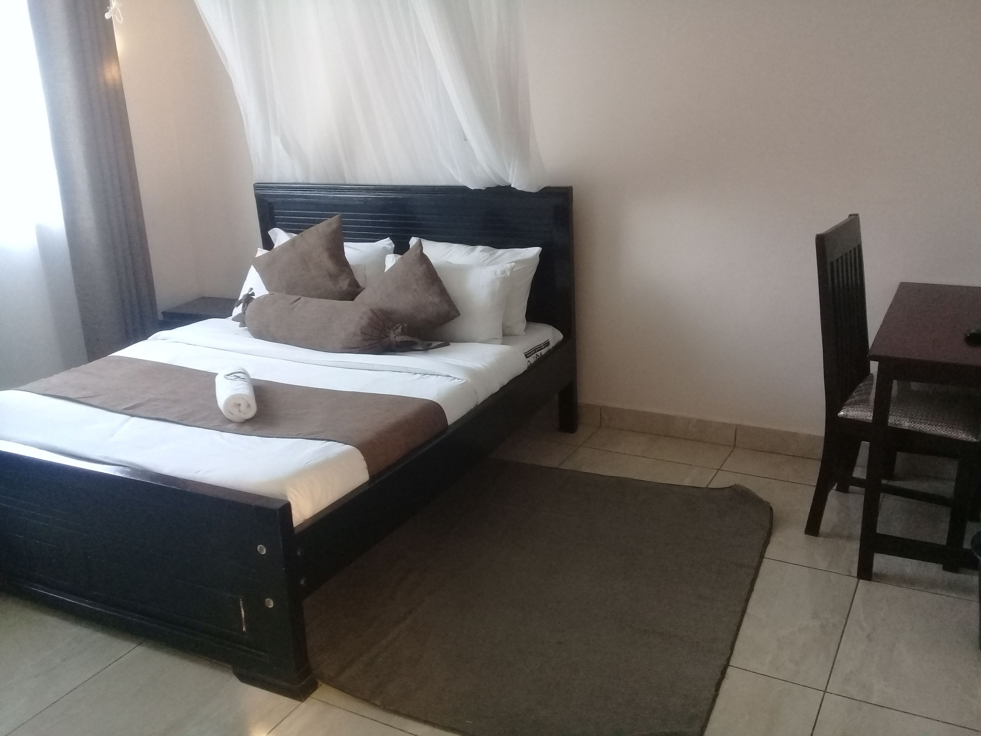 A neatly made bed with a dark headboard, a brown throw blanket, and several pillows. There is a small wooden table and a chair to the right.