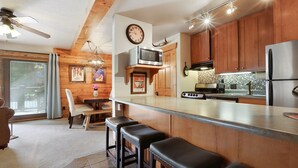Eine moderne Küche mit Holzausstattung, einem Edelstahl-Kühlschrank, einer Mikrowelle und einem Herd. Ein Essbereich mit Tisch und Stühlen sowie eine Bar mit Hockern sind ebenfalls vorhanden.