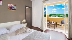 A hotel room with a bed, a nightstand, a painting, and a balcony with a view of greenery and houses. - Villa Coridigo (Tinjan)