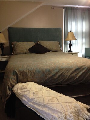 A hotel room with a bed, two bedside tables, a lamp, a chair, and a window with curtains. - Angel House B&B (Clearview)