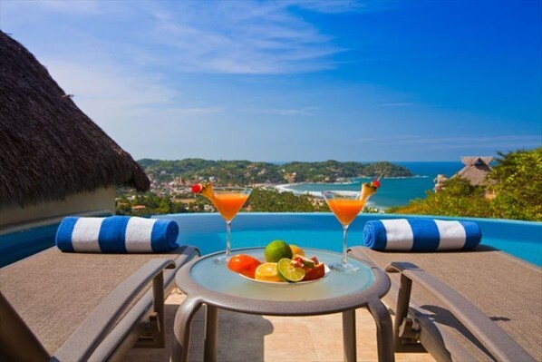A balcony with two lounge chairs, a table with a fruit plate and two drinks, and a view of the ocean and hills.