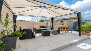 Un patio al aire libre con una zona de estar cubierta, que incluye sofás y una mesa, rodeado de plantas en macetas y una pérgola.