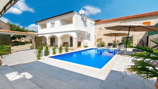 Una villa moderna con piscina, mobiliario exterior y un muro de piedra.
