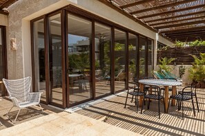 Un patio exterior cubierto con un ventanal de vidrio que da a una zona de piscina, amoblado con sillas y una mesa de metal.