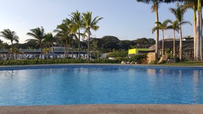A swimming pool with clear blue water, surrounded by palm trees and a building with a yellow facade.