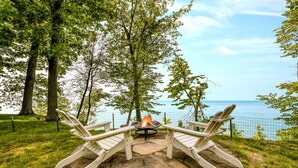 A stone fire pit with two Adirondack chairs facing it, surrounded by a grassy area with trees and a view of the water. - Blue Water Retreat - Spacious Lake Michigan retreat with In-ground pool (Benton Harbor)