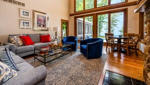 A spacious living room with a gray sofa, red cushions, a coffee table, and blue armchairs. There are framed pictures on the wall, a fireplace, and large windows offering a view of the outdoors. - Blue Water Retreat - Spacious Lake Michigan retreat with In-ground pool (Benton Harbor)