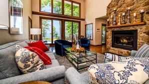 A spacious living room with a stone fireplace, large windows offering a view of the outdoors, and a cozy seating area with patterned cushions. - Blue Water Retreat - Spacious Lake Michigan retreat with In-ground pool (Benton Harbor)