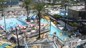 Una amplia piscina exterior con varias pistas, rodeada de palmeras y sombrillas.