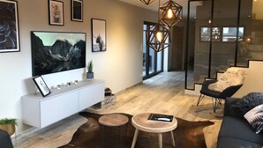 Uma sala de estar moderna com uma grande televisão de tela plana, um console branco, uma mesa de centro redonda de madeira, um sofá de couro preto e uma poltrona preta, além de um painel divisório de vidro.