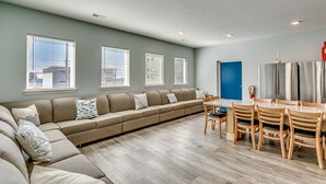 A modern living room with a sectional sofa, dining table, and chairs. There are large windows with blinds, a blue door, and a stainless steel refrigerator. - ACROSS FROM the BEACH SLEEPS 50, HUGE POOL+SPA, CHEF'S KITCHEN, GAME+MEDIA ROOM (North Myrtle Beach)