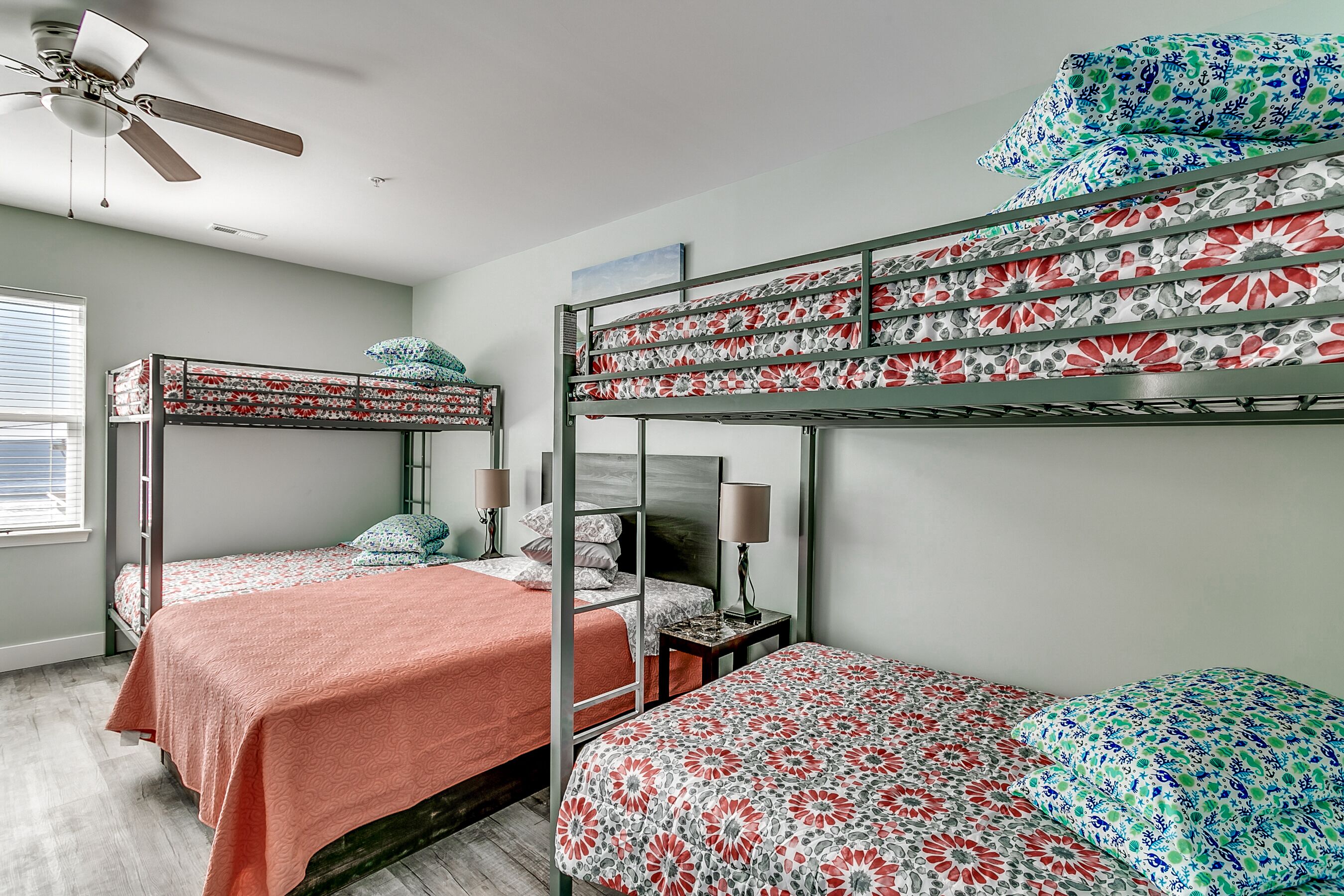 Une chambre avec un lit superposé, deux lits, un ventilateur de plafond et une fenêtre avec des stores.