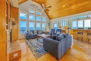 A spacious living room with a large gray sofa, a coffee table, and a wooden ceiling fan. There are large windows offering a view of the ocean.