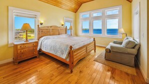 Une chambre avec un lit en bois, deux lampes de chevet, une commode en bois, un canapé gris et de grandes fenêtres donnant sur la mer.