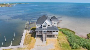 Une maison à deux étages avec une jetée qui s’avance dans l’eau, entourée d’une pelouse et d’une plage de sable.