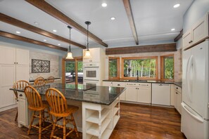 A spacious kitchen with a large island, wooden chairs, white cabinets, and a view of the outdoors through large windows.