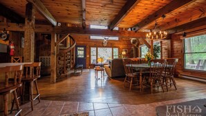 Interior rústico de uma cabana com sala de jantar, escada de madeira e janelas amplas.