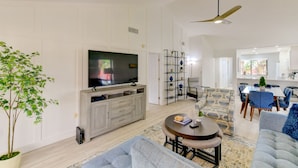 A spacious living room with a large flat-screen TV, a grey sofa, a coffee table, and a dining area with a table and chairs.