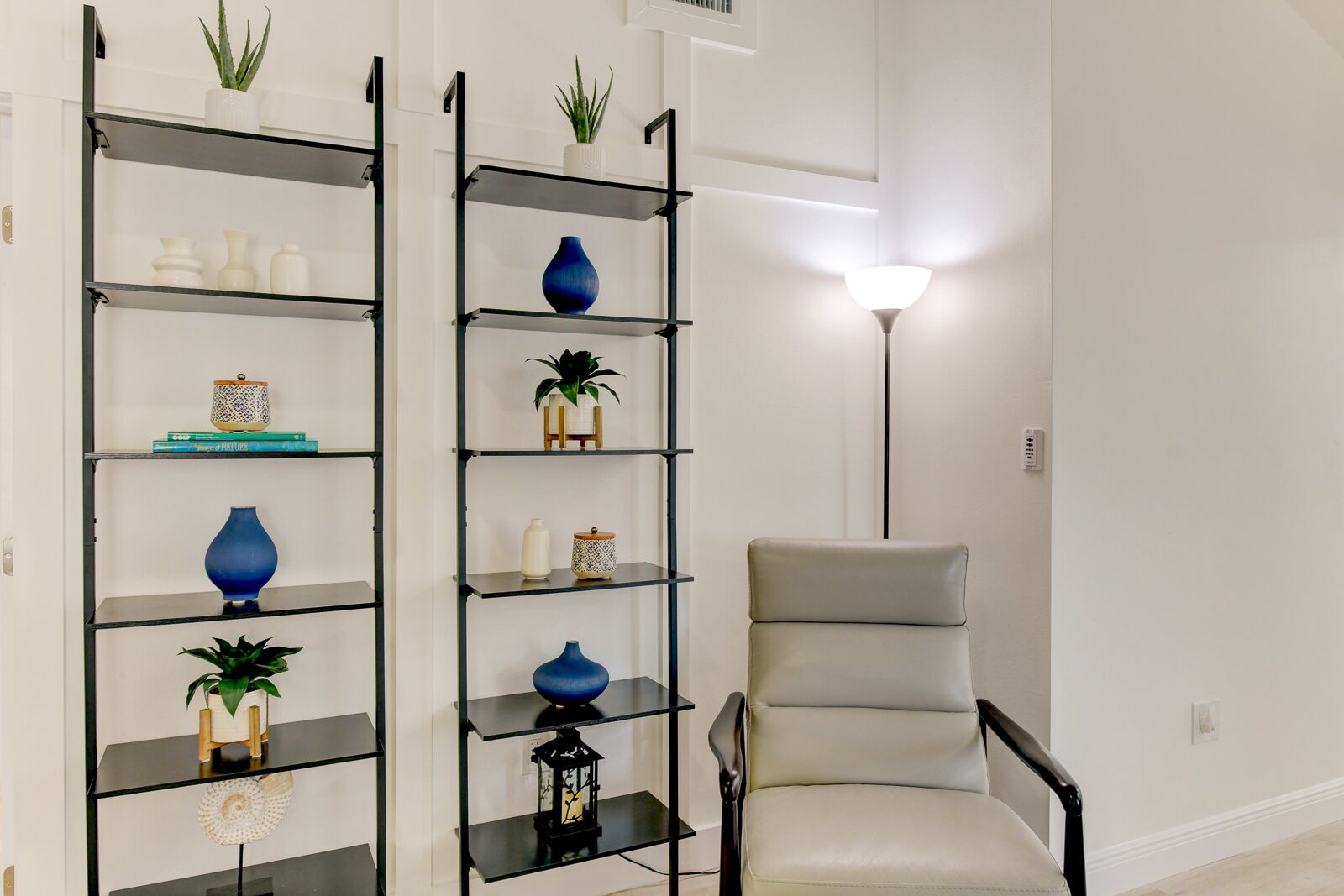 Ein Wohnzimmer mit einem beigen Sessel, einem schwarzen Metallregal mit Dekoration und einer Stehlampe.
