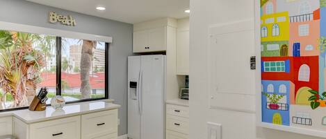 A kitchen with white cabinets, a white refrigerator, and a window with a view of palm trees and buildings.