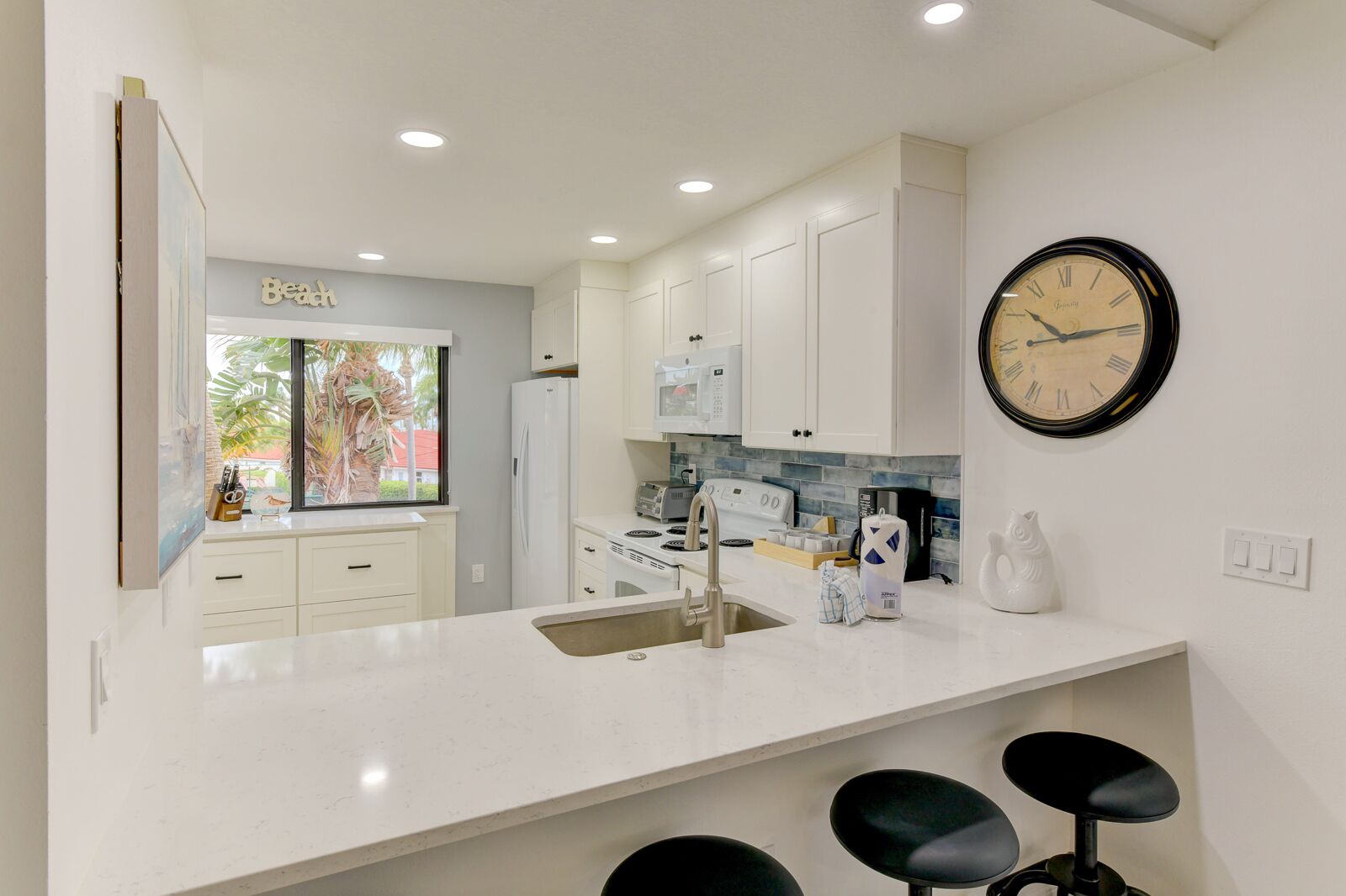 A modern kitchen with white cabinets, a large window with a view of palm trees, a black clock, and a sink.