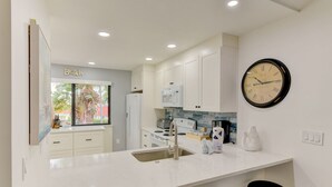 A modern kitchen with white cabinets, a large window with a view of palm trees, a black clock, and a sink.