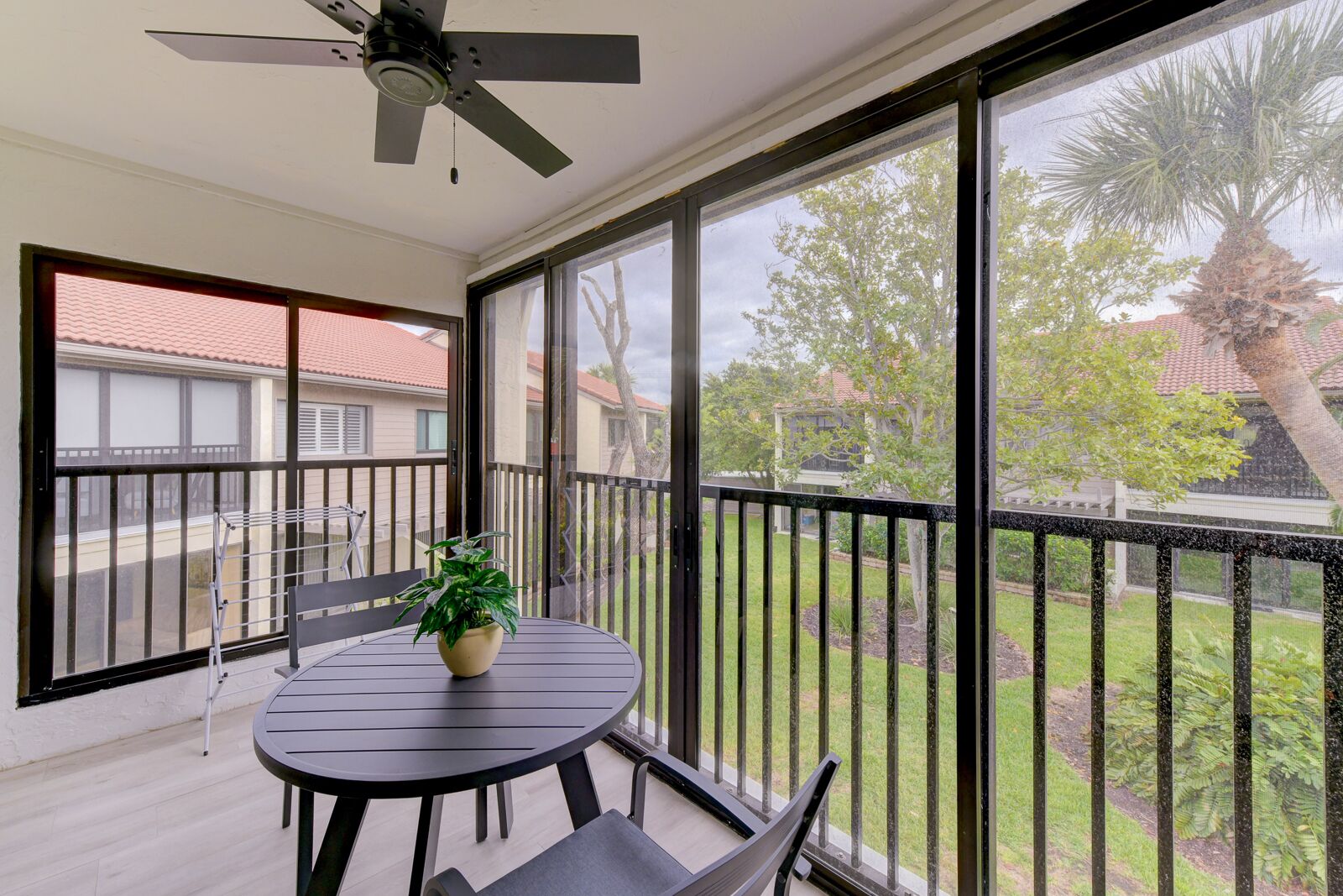 Uma varanda com mesa e cadeiras, um vaso com planta e vista para uma área residencial com casas e árvores.