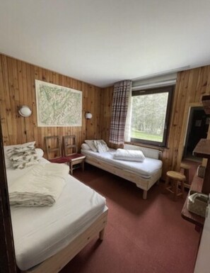Ein Zimmer mit einem großen Bett, einem kleineren Bett, einem Sessel, einem Tisch und einem fensterbelogenen Fenster.