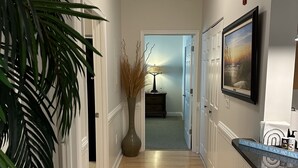 A hallway with wooden flooring, white wainscoting, a large vase with dried plants, a framed picture on the wall, and a door leading to another room. - 2 Bedrooms with king beds and private bathroom for both. (Charleston)