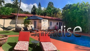 A poolside area with lounge chairs and a small table, surrounded by artificial grass and a red pathway.