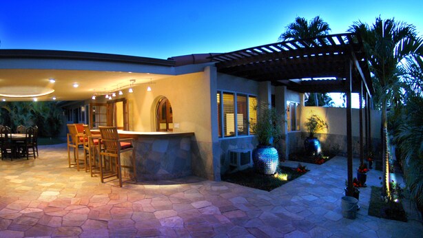 A modern outdoor patio with a stone floor, a dining area with chairs and a table, and a bar area with a counter and stools.