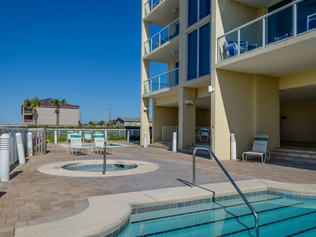 A hotel pool area with a circular hot tub, lounge chairs, and a multi-story building with balconies.