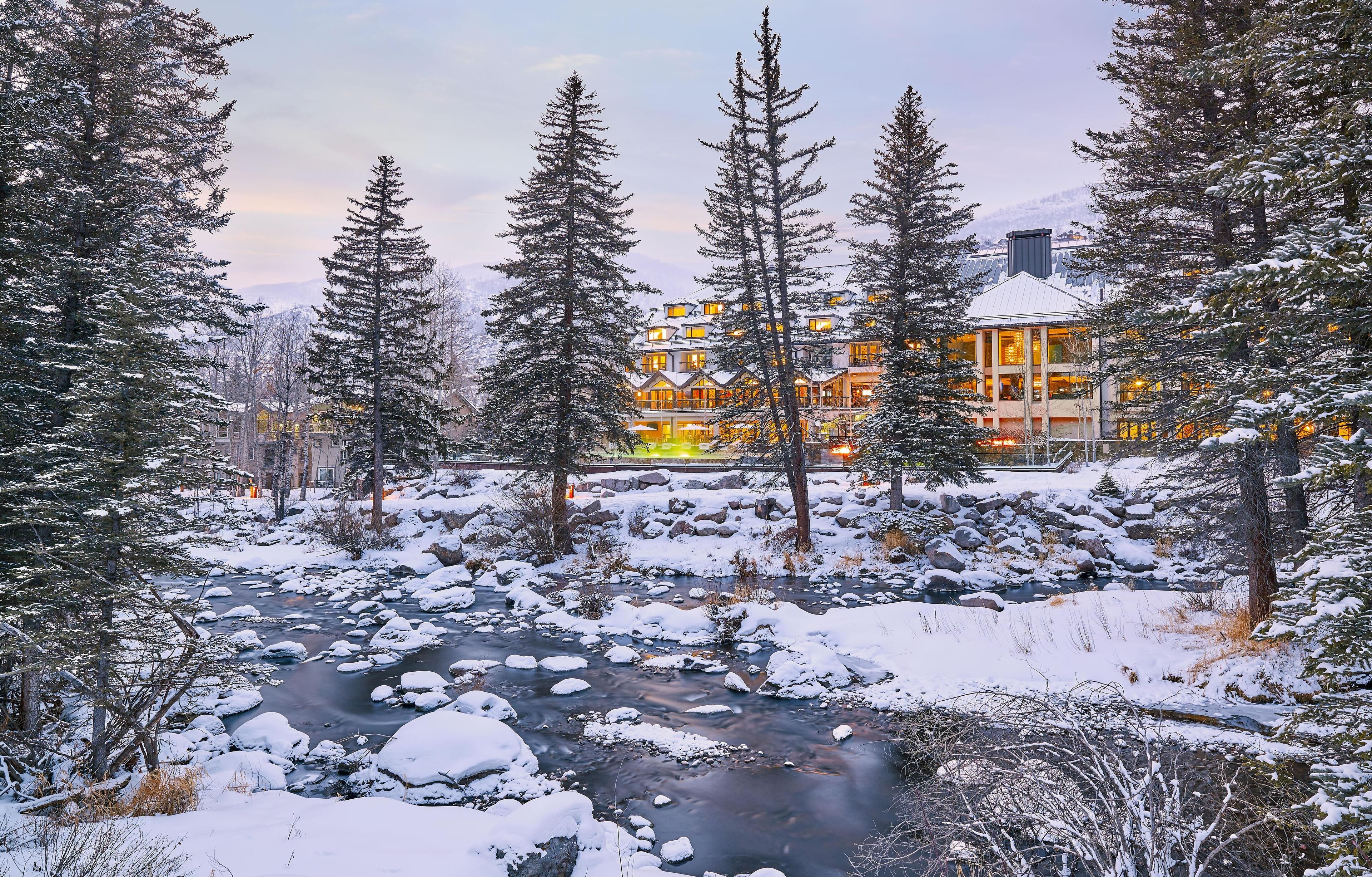 A lodge with a river in the foreground, surrounded by snow-covered trees.