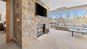 A living room with a stone fireplace, a flat-screen TV, a sofa, and large windows. - Mountain-View Penthouse w/ convenient access to Grand Hyatt Vail (Vail)