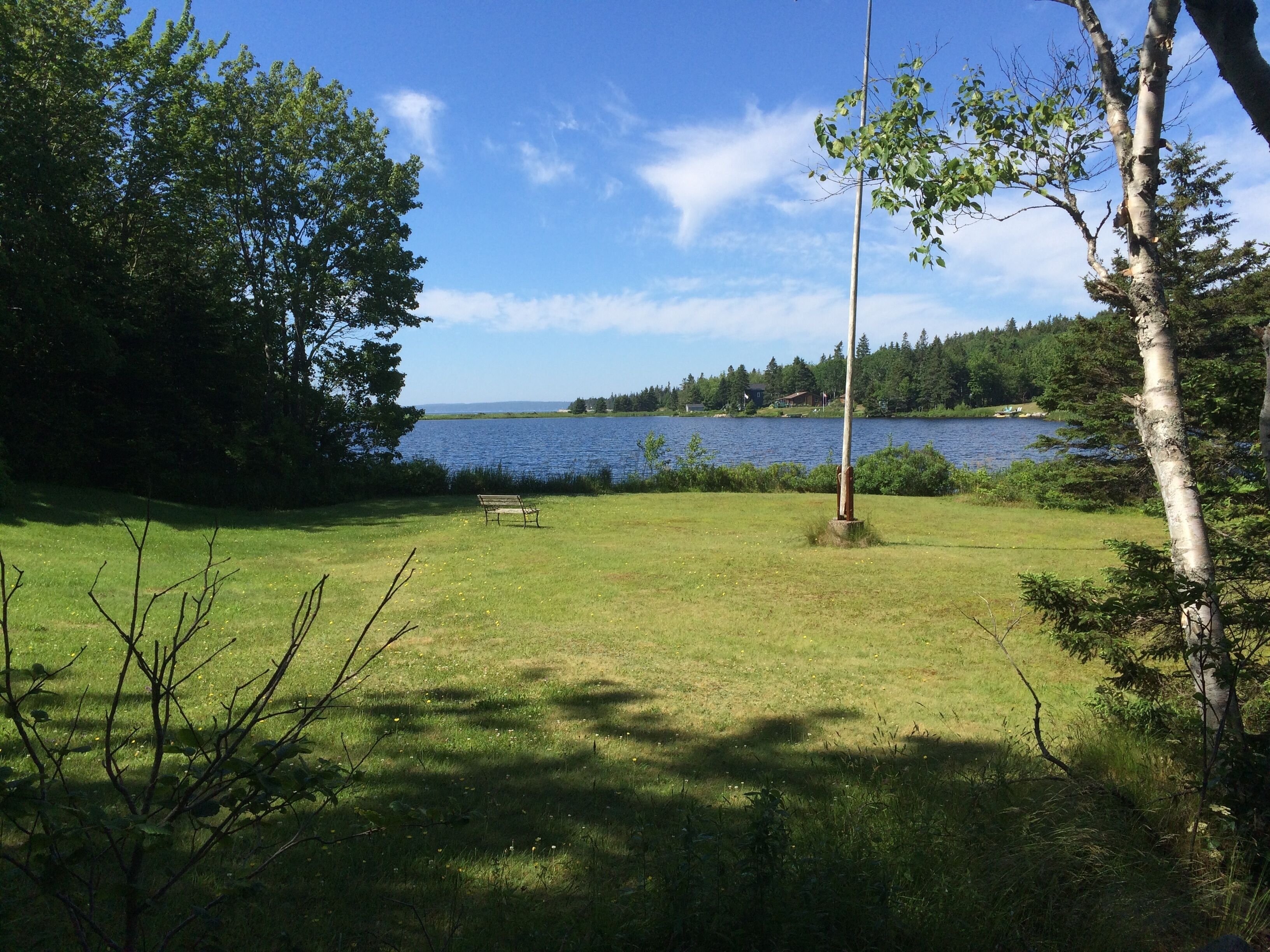 Un champ verdoyant avec un banc et un mât de drapeau, d’où l’on a une vue sur un lac, avec des arbres et des maisons en arrière-plan.