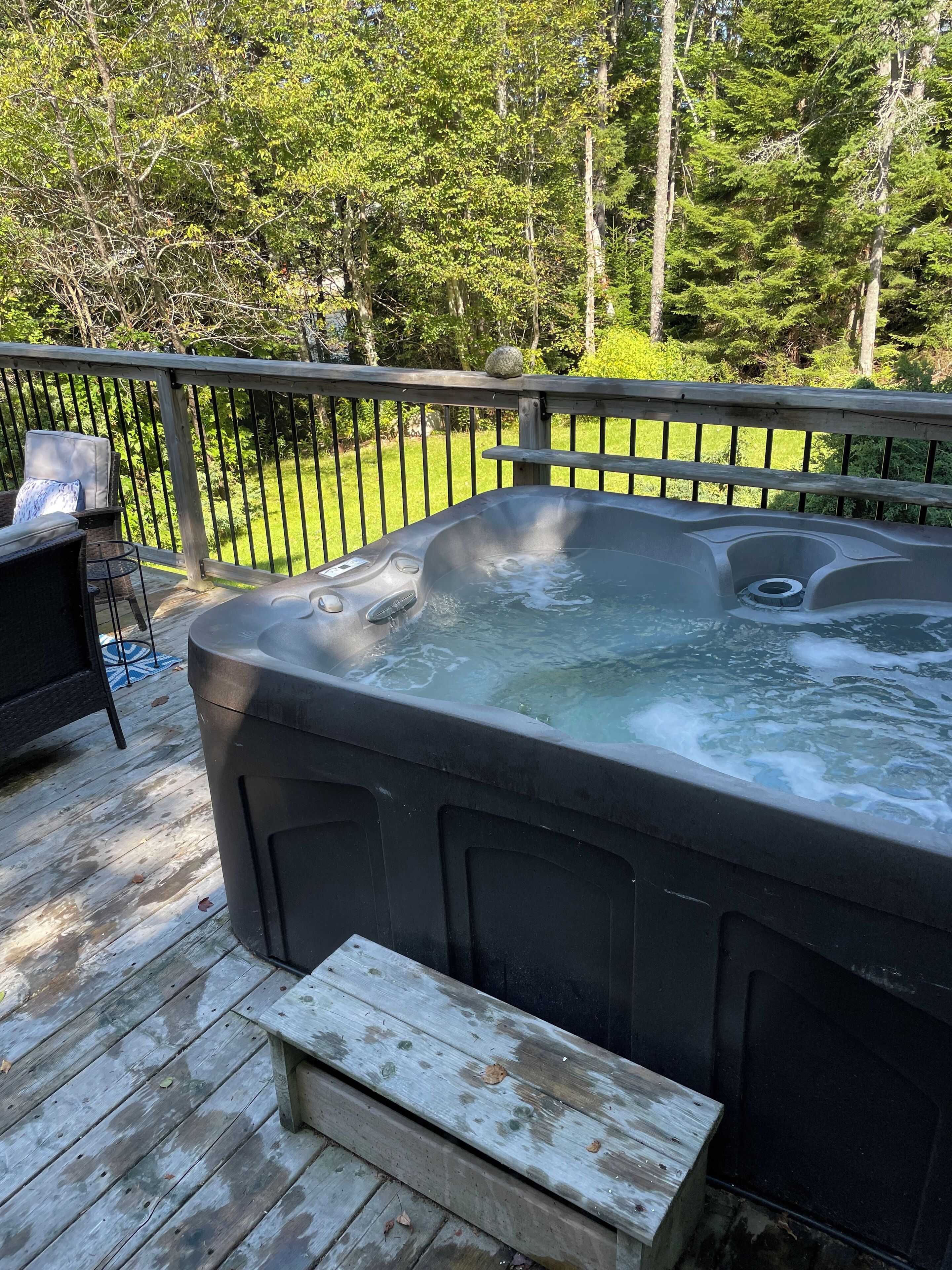 A hot tub on a wooden deck with a bench, surrounded by a metal fence and greenery.