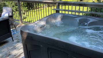 A hot tub on a wooden deck with a bench, surrounded by a metal fence and greenery.