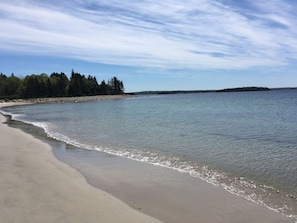 A beach with clear water, a sandy shore, and a forested island in the distance.