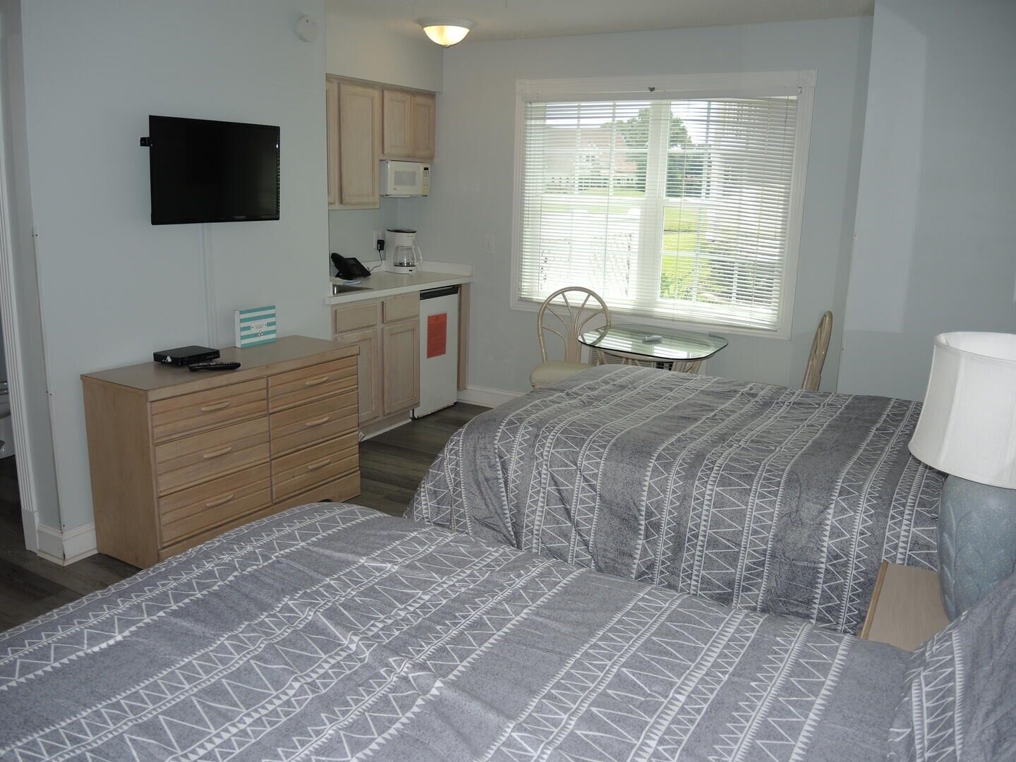 A bedroom with two beds, a desk, a chair, a TV, a microwave, and a window with blinds.