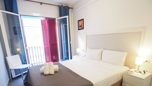 A hotel room with a bed, a bedside table with a lamp, a chair, and a view of the outside through a window with red shutters. - Balcony View Apartment by Hello Homes Sitges (Sitges)